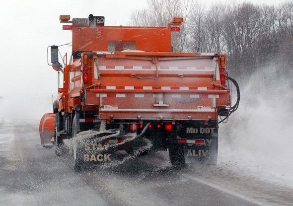 A Minnesota Department of Transportation orange snowplow clearing a snowy road, with visible spray of salt and snow from the rear. Sign on the back advises to 