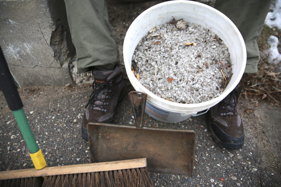 A person wearing winter boots sits next to a sidewalk with a white five-gallon bucket filled with salt granules and debris on it. A broom and a dustpan are resting against the bucket.