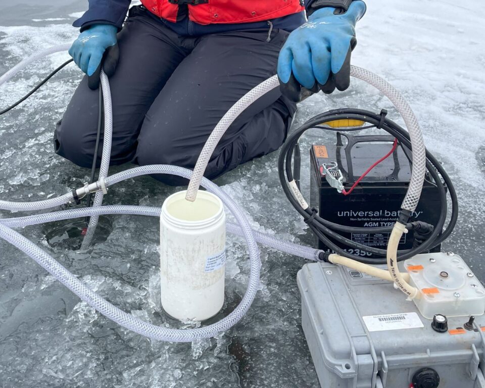 A person kneels next to a hole drilled in a frozen lake. They are holding a plastic tube that is bringing water from the hole to a plastic sample container via a battery powered pump. 