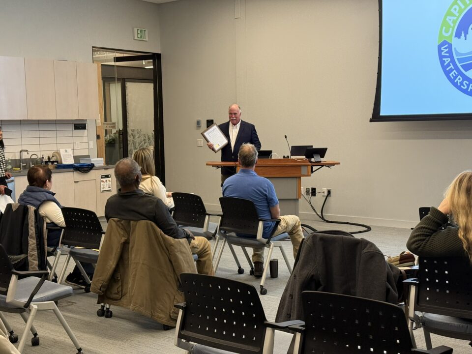 A person holding a framed proclamation stands at a podium in a meeting room with an audience seated facing them.