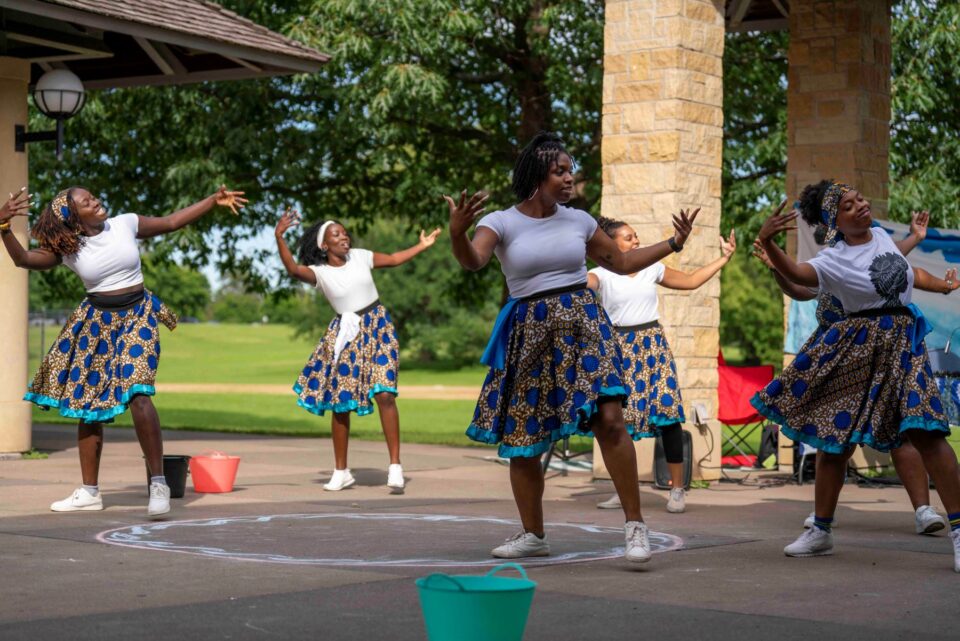 Six dancers in matching blue and yellow African print skirts and white shirts perform outdoors. They gracefully open their arms with elbows bent as they step and look to the side.