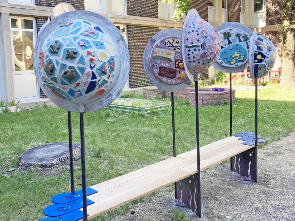 Colorful tile mosaics with turtles, fish, raindrops, a tree-lined pond with ducks and assorted shapes cover several half sphere bubbles on poles rising from a slatted bench along a grassy space next to a brick school building. 