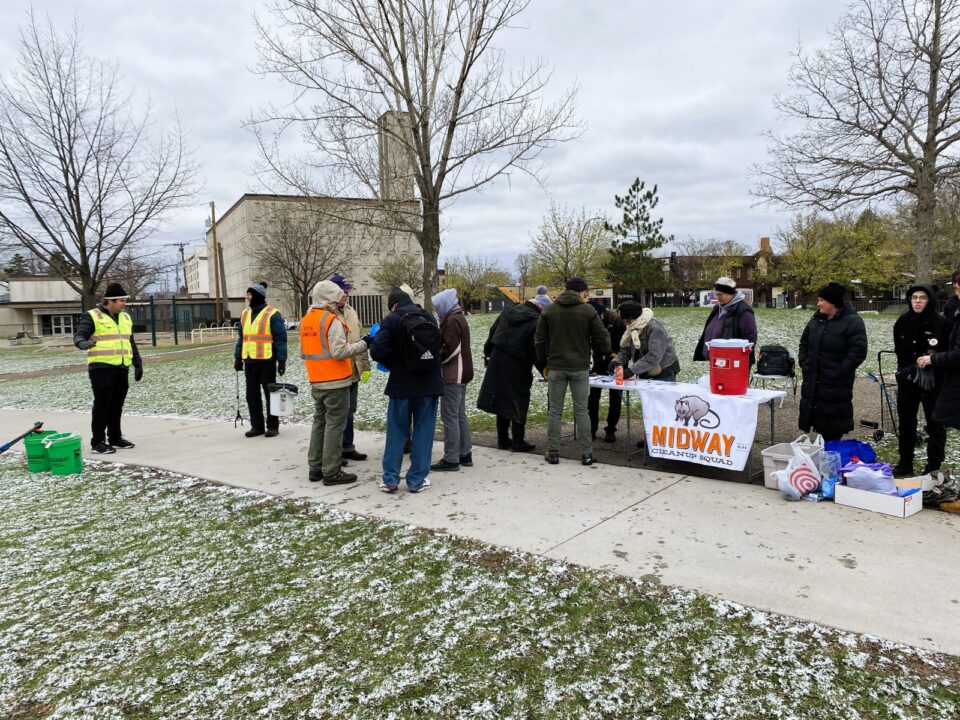  Several people stand around a table with a sign featuring an illustrated opossum and the words “Midway Cleanup Squad” along a pathway in a snow dusted park.