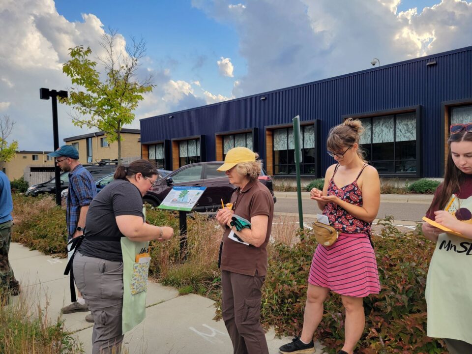 Several people stand on a sidewalk with native grass and shrubs on both sides, next to a small parking lot in front of a blue-clad modern office building. 