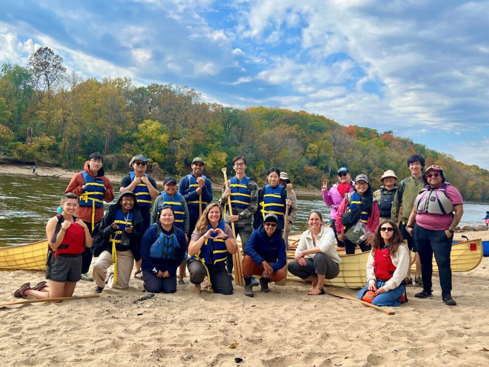  A large group of smiling people wearing life vests pose for a photo on a sandy beach with canoes and paddles around them. The Mississippi River in the background under a blue sky streaked with clouds. 
