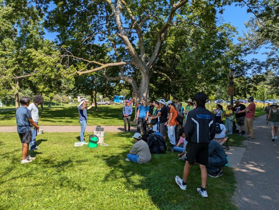 A group of teenagers standing under trees in a grassy park space in dappled shade. They are listening to someone speaking next to water quality monitoring equipment. 