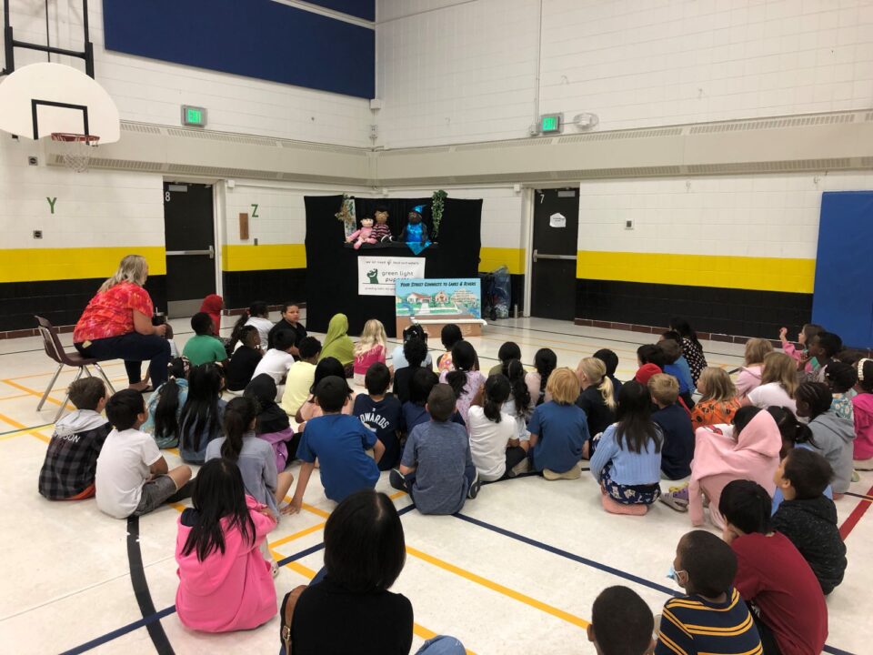 A large group of children sitting in a school gym, attentively watching a puppet show. A sign in front of the puppet stage reads “Your street connects to lakes and rivers.”