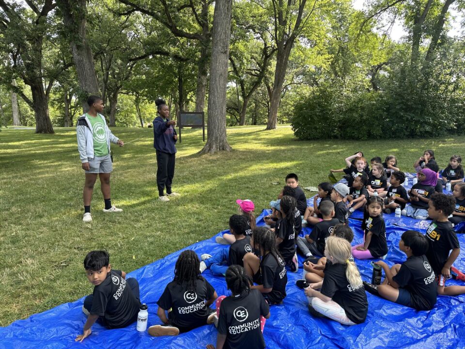 Two instructors leading a group of children seated on a blue tarp in a grassy area of a park surrounded by trees.  