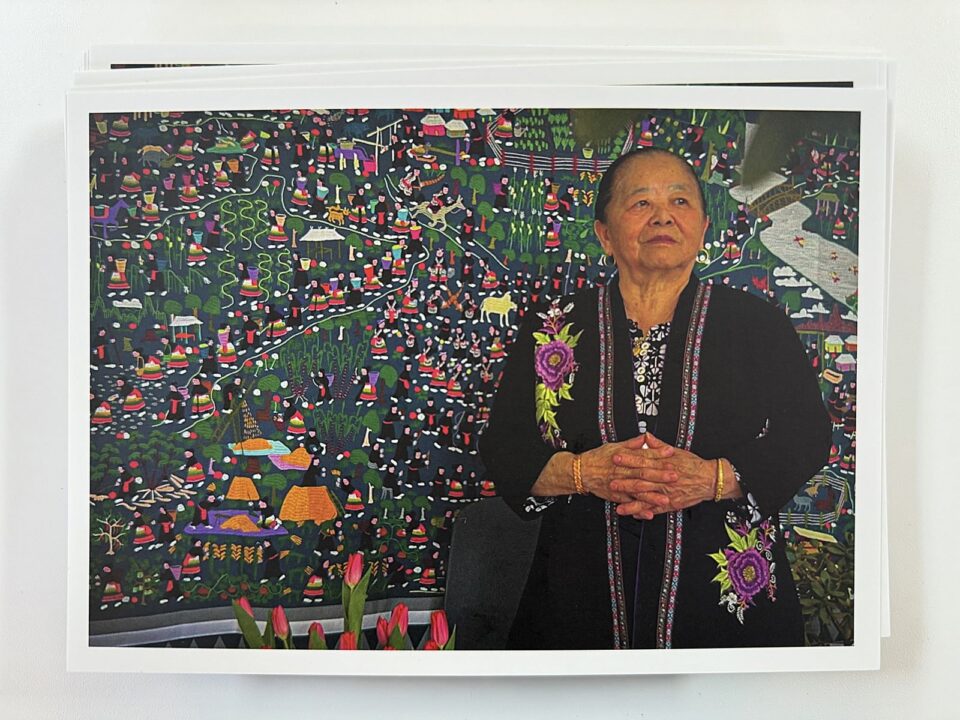 Chia Xiong standing in front of a large, colorful quilt cloth that show many people farming, working and dancing in detailed scenes. She is wearing a long black jacket with a floral ribboned border and large purple flowers surrounded by green leaves. 