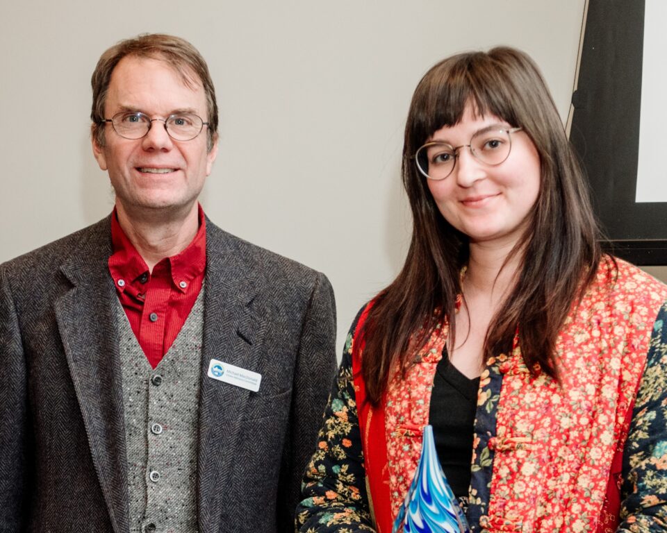 Two smiling people in a conference room. Mike MacDonald is on the left, wearing a snazzy red shirt under a gray suit jacket and vest. Erin Spry is on the right wearing a colorful patterned jacket. 