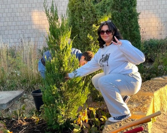 A smiling person squatting on a low stone wall while holding up two fingers in a peace sign with one hand and holding an evergreen shrub with their other hand as another person plants it. 