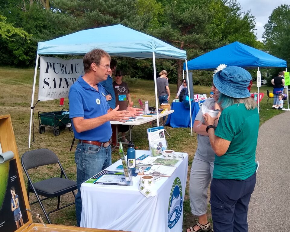 Mike MacDonald standing and talking outdoors to two people across a table with a tablecloth showing Capitol Region Watershed District’s logo on the front and covered with flyers and promotional materials. In the background there are popup canopy tents with tables in a grassy area along a paved path. 