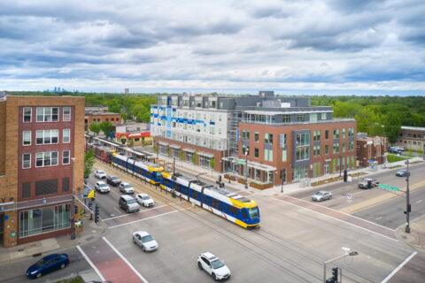 Aerial view of a busy urban intersection with a light rail train, cars driving on the street, and modern five-story building centered in the frame on an overcast day.