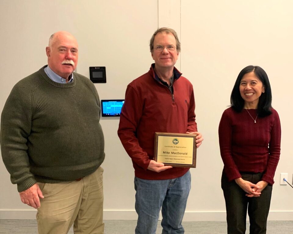 Three smiling people standing in a meeting room. The man in the center is holding a recognition plaque with Capitol Region Watershed District’s logo and the name “Mike MacDonald”. 