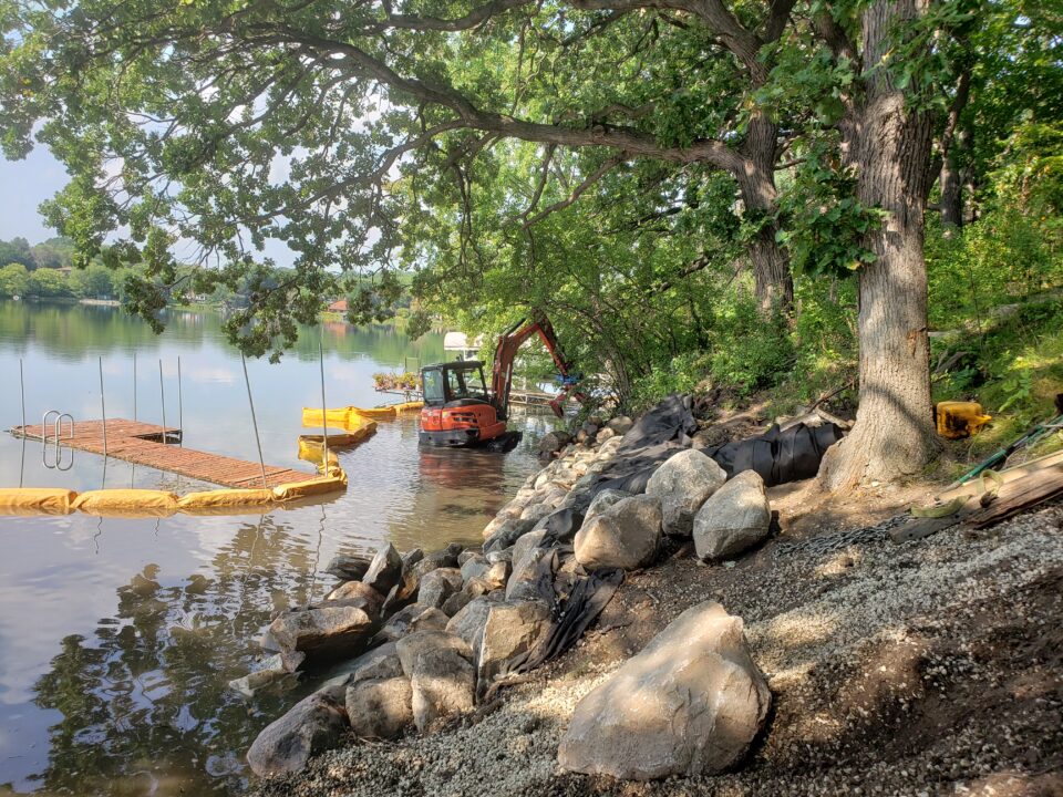 Gravel and large rocks piled on a shade-dappled lakeshore. In the lake, a floating yellow containment boom is running parallel to shore, with a wooden dock on the outside of the boom. An construction vehicle with a claw to move the rocks is sitting in shallow water in the near side of the boom. 