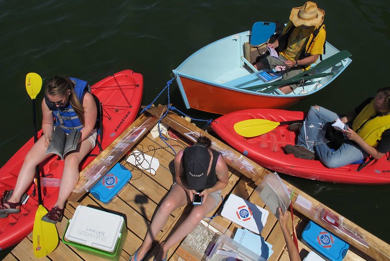 An overhead view of someone sitting on a floating wooden platform on a lake with blue and white plastic bins, a cooler, and stacks of booklets in the center. The outer edges of the platform have angled wooden shelves holding magazines and books inside plastic covers. Two red kayaks and a light blue and orange rowboat are tied to the platform while people sitting in them are reading books and magazines.