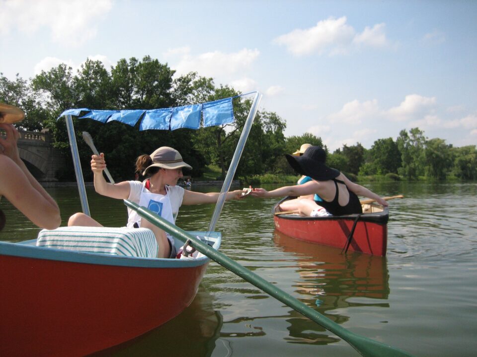 A person wearing a sunhat and apron sits in a cheery orange and blue rowboat with blue ice cream cone printed flag bunting extending above them while reaching back to hand money to a person in a black sunhat and swimsuit in a red and black canoe. The boats are on a calm lake surrounded by green trees with an arched stonework bridge and a low stone wall along one side, under a sunny blue sky with soft white clouds. 