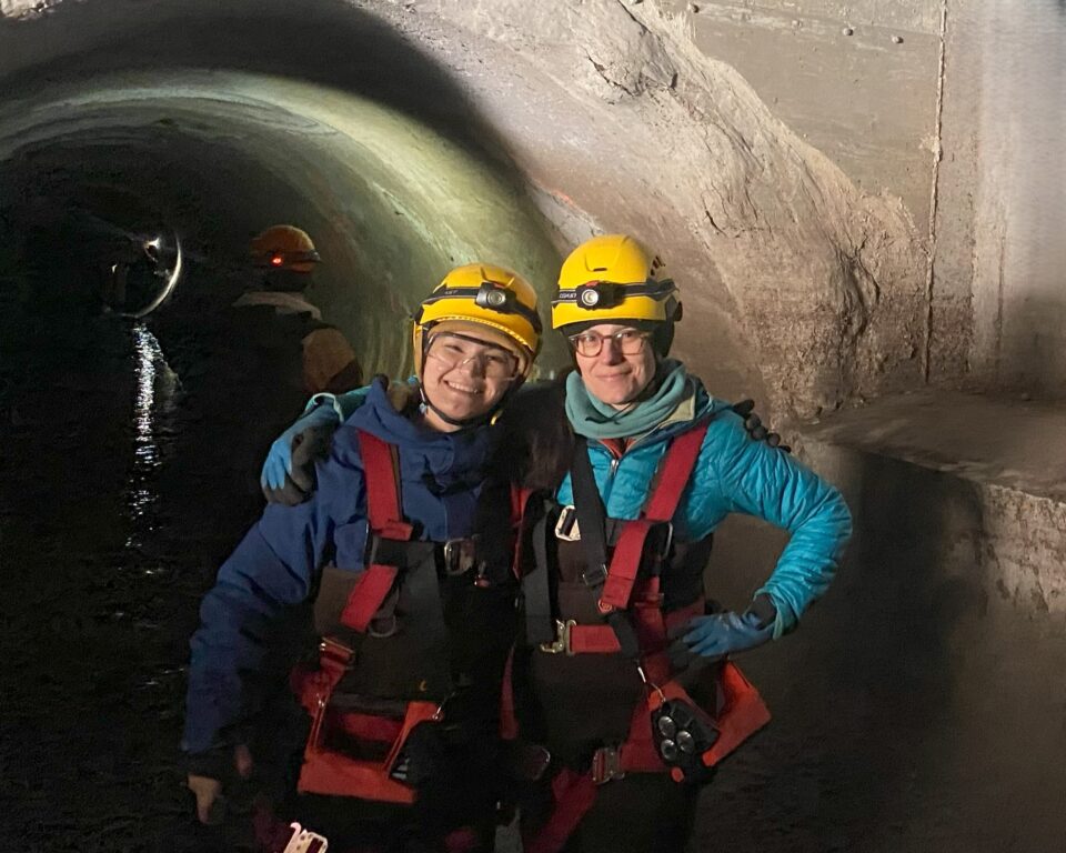 Two smiling women standing in a dimly lit concrete tunnel. They are wearing yellow helmets with headlamps, and safety glasses. They are wearing red safety harnesses over insulated jackets and brown chest waders. 