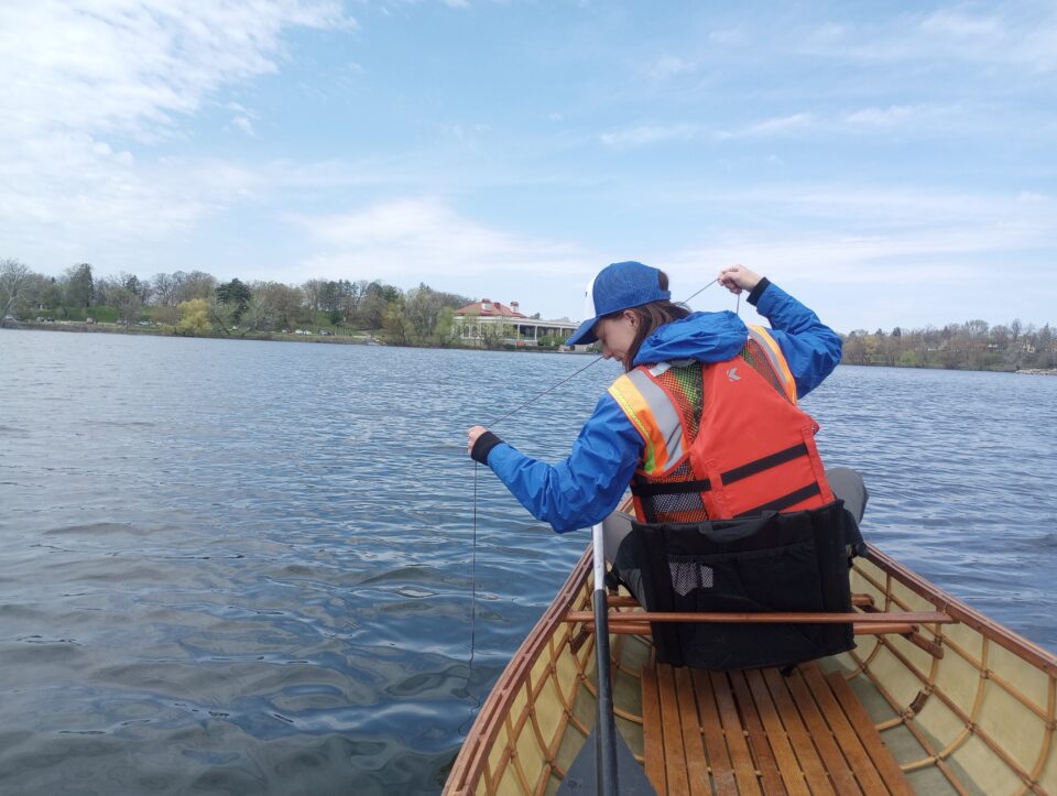 A person in a life jacket and hat sitting in a canoe in Como Lake. They are holding a line of string and looking excitedly down at the water.