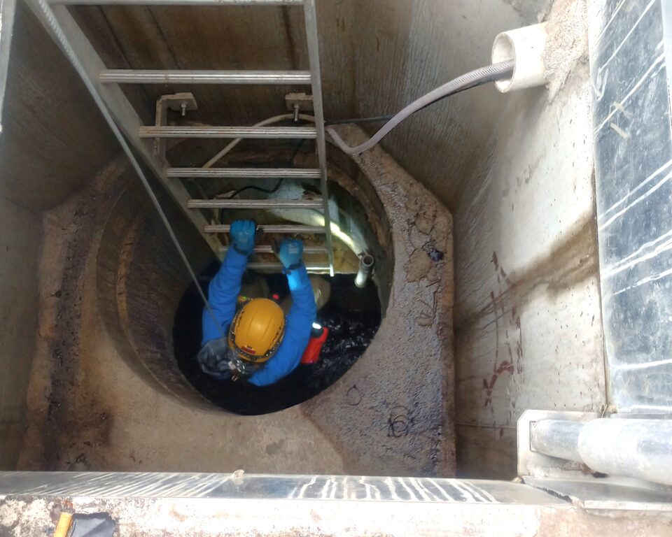 Person in a safety helmet and gloves climbing down a ladder inside a concrete pipe with water in the bottom. 