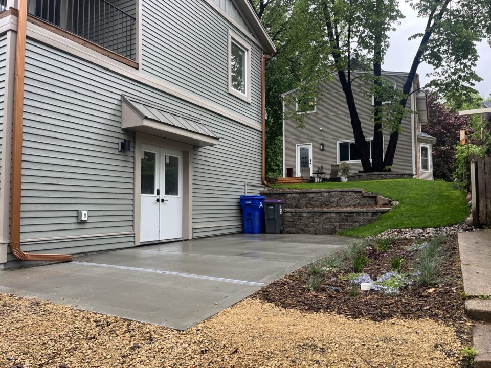 A downspout of a house leads to a long metal grate in a concrete slab. The grate connects the downspout to a mulched rain garden.  