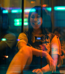 A laughing woman with dark hair and hip clothing casually sits behind window reflecting glowing lights. 