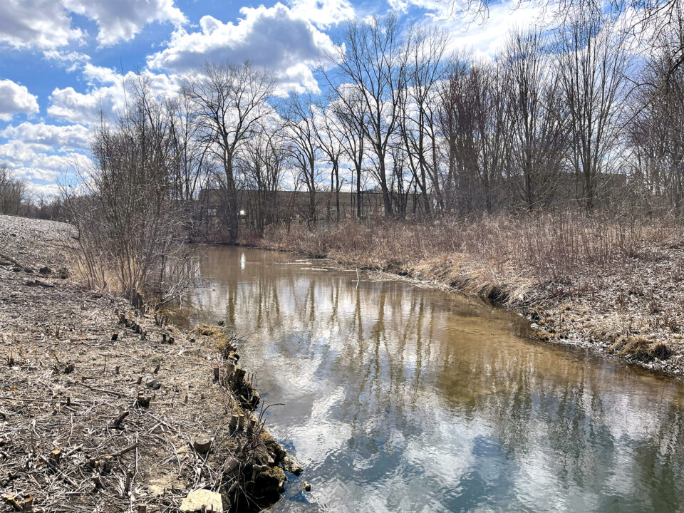 A shallow stream surrounded by brown grass and bare trees on both sides with an industrial building in the background.