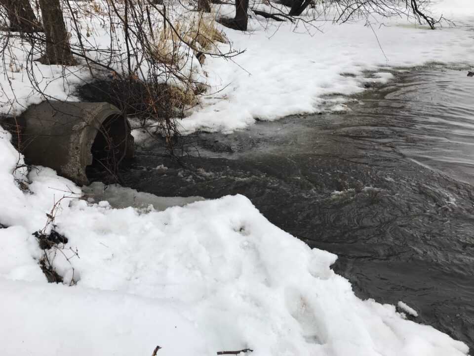Water flowing from a concrete culvert into lake with snow-covered shores.  