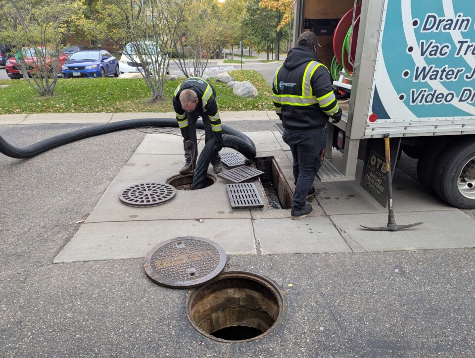 Two workers near a large truck work in a grate and manholes in a parking lot with the covers removed. One worker holds a large, flexible tube going into one of the open manholes.