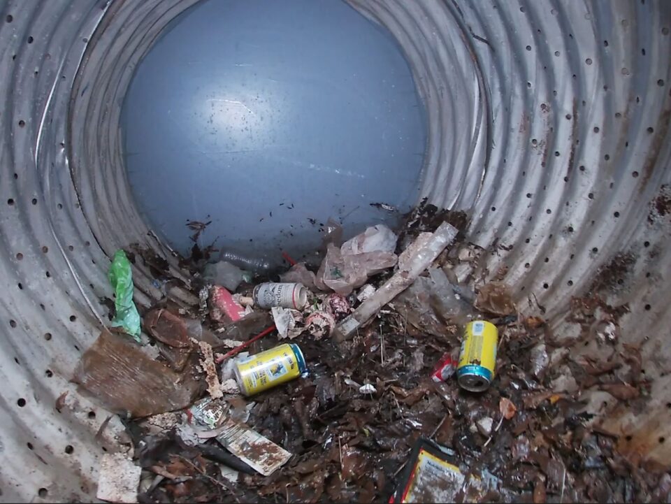 Trash, leaves and debris sit in the bottom of a perforated metal pipe. 