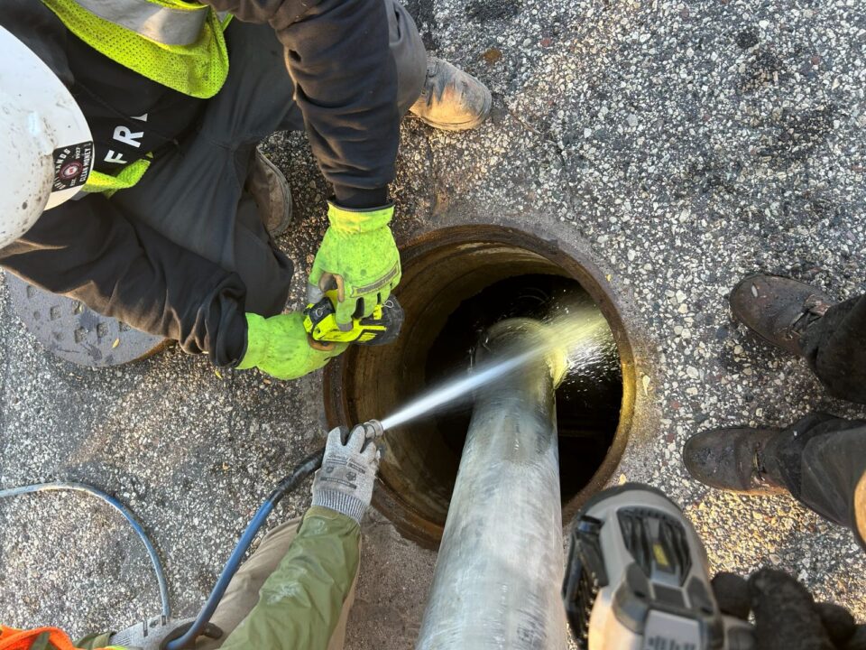 An open manhole in a parking lot has a metal tube in it. A worker wearing work gloves holds a hose spraying into the manhole while another worker holds a light on the stream of spray. 