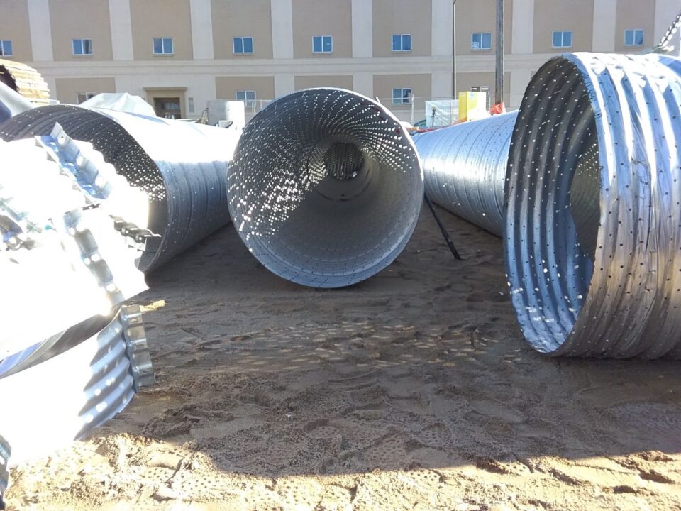 Dots of light shine through a large, corrugated metal pipe perforated with small holes sitting on smooth sand in a construction site.