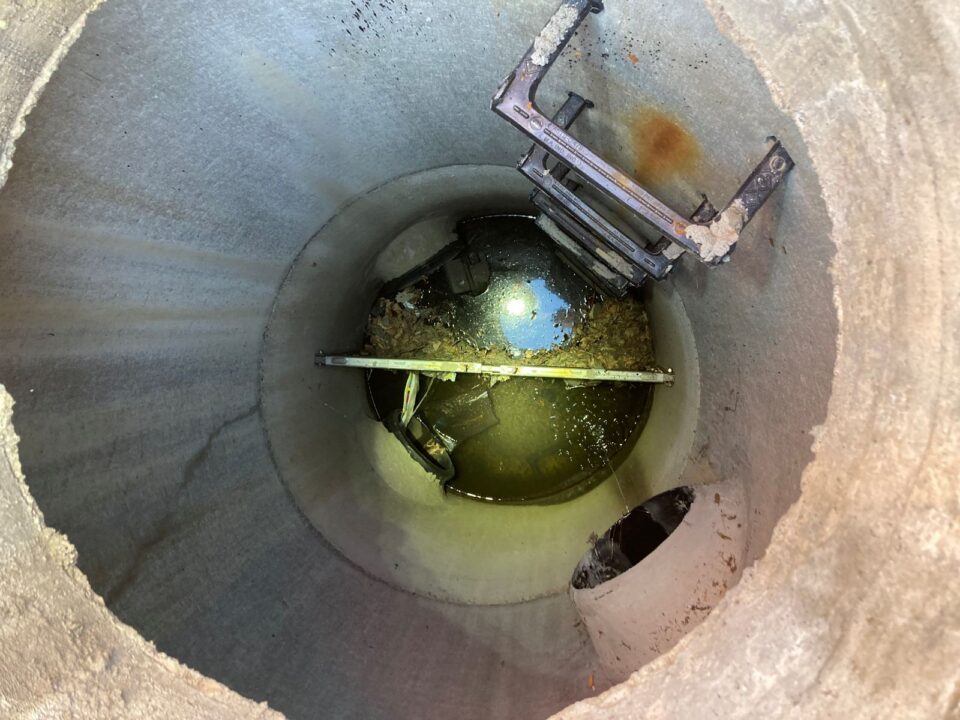 A concrete pipe with an access ladder on the side has standing water at the bottom of it with floating trash. A circle of bright light and blue sky from above are reflected in the water. 