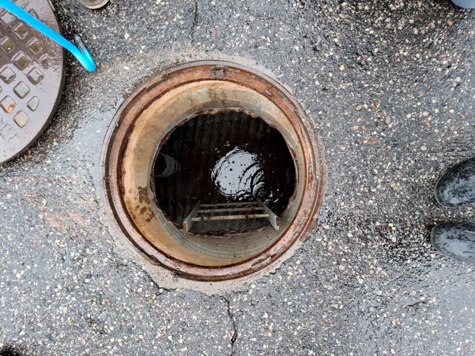An open manhole with an access ladder in a parking lot wet with rain has standing water at the bottom of it. Light from above is reflected in the water with raindrops breaking the surface. 