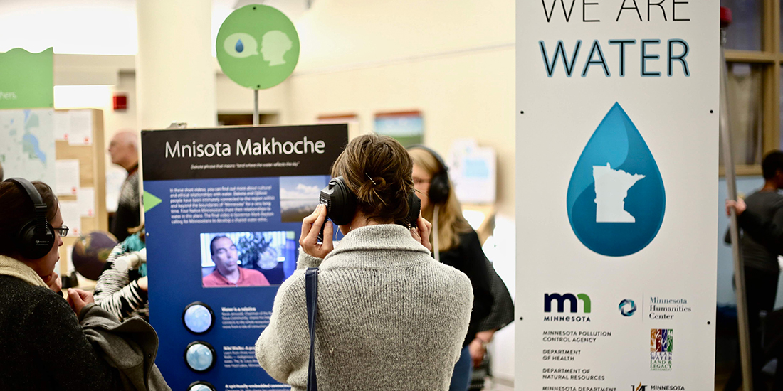People wearing headphones engage with exhibits at an event focused on water and culture in Minnesota, featuring the Minnesota Humanities Center and the Minnesota Pollution Control Agency. Signs display information about Mnisota Makhoche, with images and text.