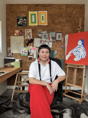 Christina Vang sitting in an office chair in an art studio. Behind her is a cork wall pinned with colorful pictures, and an easel with a painting.