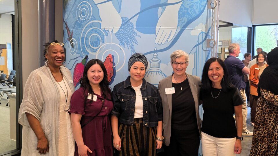 Five smiling women standing in front of a water-themed mural on a large cistern.