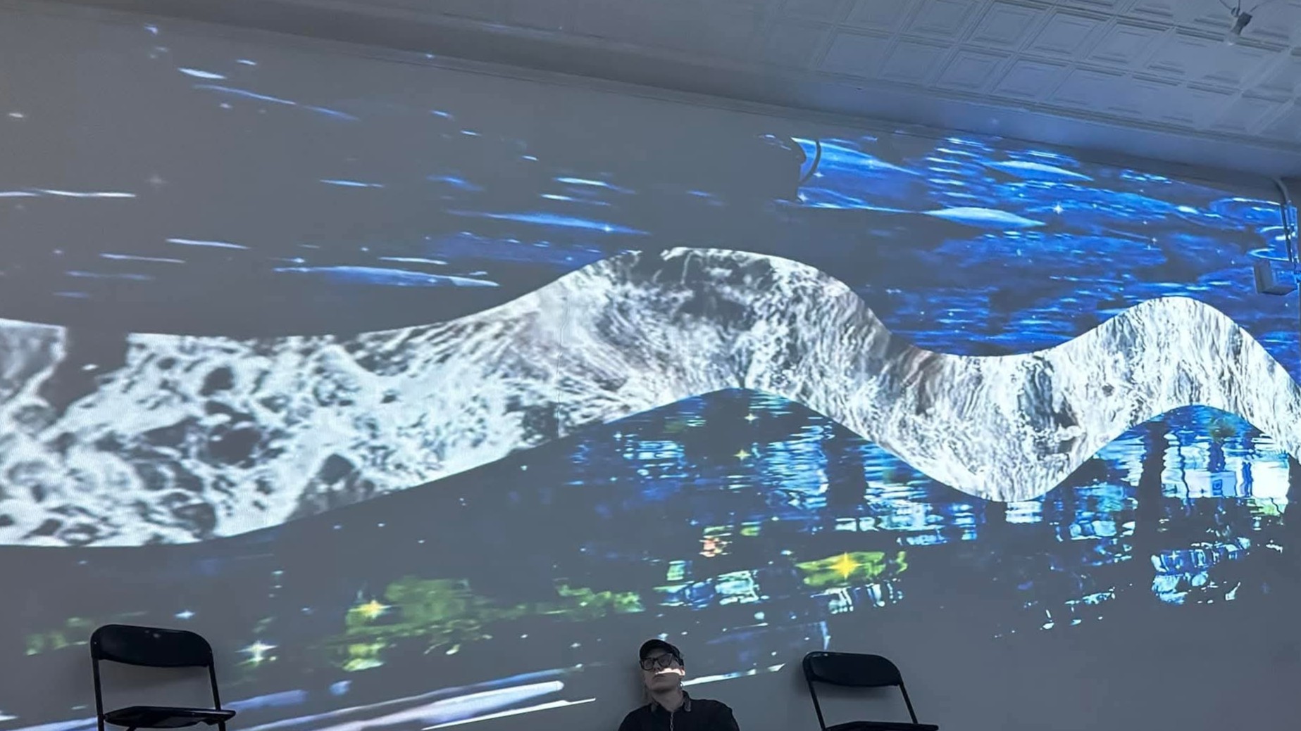 A wavy, collaged projection on a wall with a person sitting below looking up.