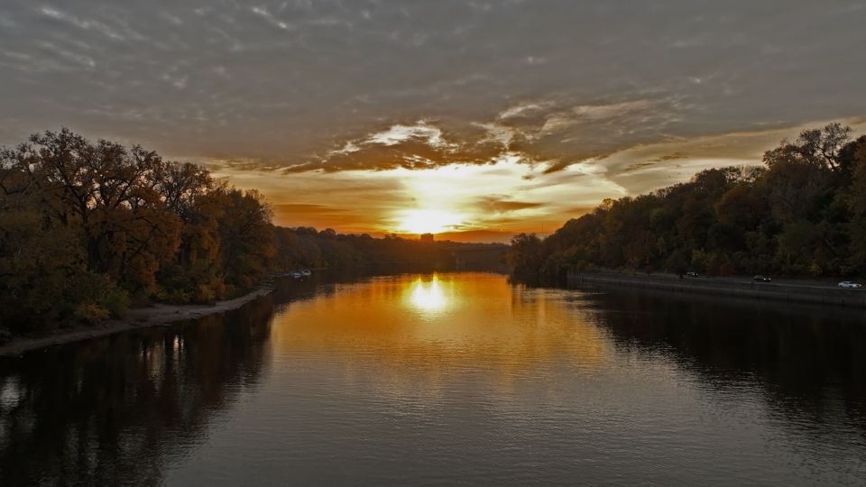 Mississippi River at sunset with the low sun and tree-lined banks reflecting off the water. An elevated road with cars is on one side of the river and a sandy beach is on the other.
