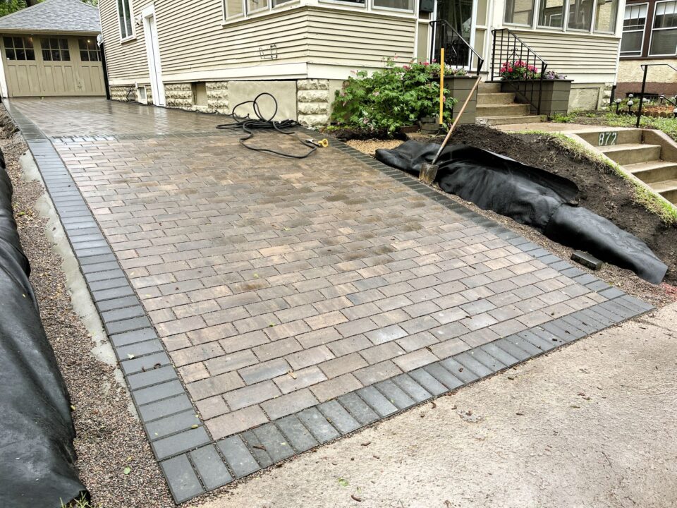 A newly paved driveway made of brown bricks with a border of darker bricks leads along the side of a house. A garage is visible in the background. On the right side of the driveway, in front of the house, there is landscaping work in progress with black tarp and exposed soil.