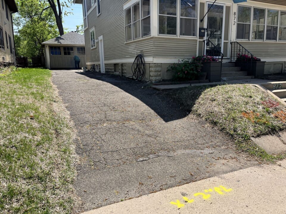 An old asphalt-paved driveway leads along the side of a house with beige siding and a large porch. A garage is visible in the background. Yellow utility markings are on the sidewalk in front of the driveway. 