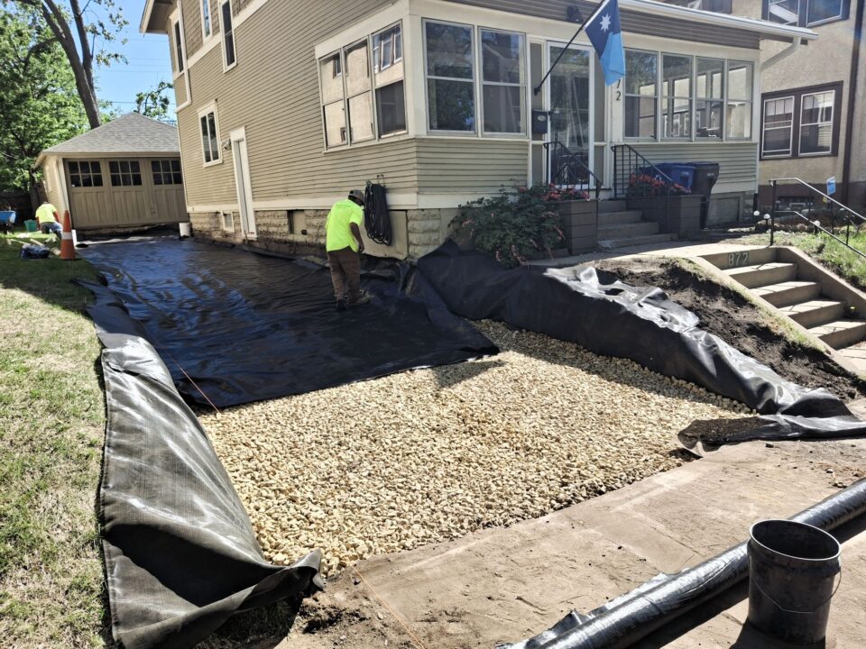 A person working on a driveway project outside a house is laying black landscaping fabric over gravel on top of another layer of black fabric. A garage is visible in the background.