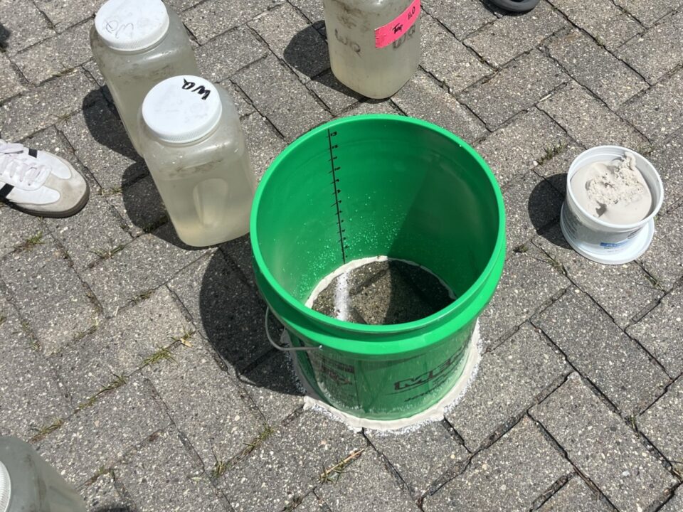 A green bottomless 5-gallon bucket with measure markings on the inside is puttied to a brick-paved lot. The bucket is partially filled with water. The bucket is surrounded by several clear plastic containers of water.