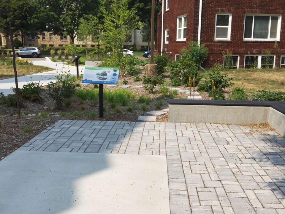 A brick-paved patio surrounded by landscaped space with paved walkways and a rain garden featuring an informational sign. A red brick building stands in the background.