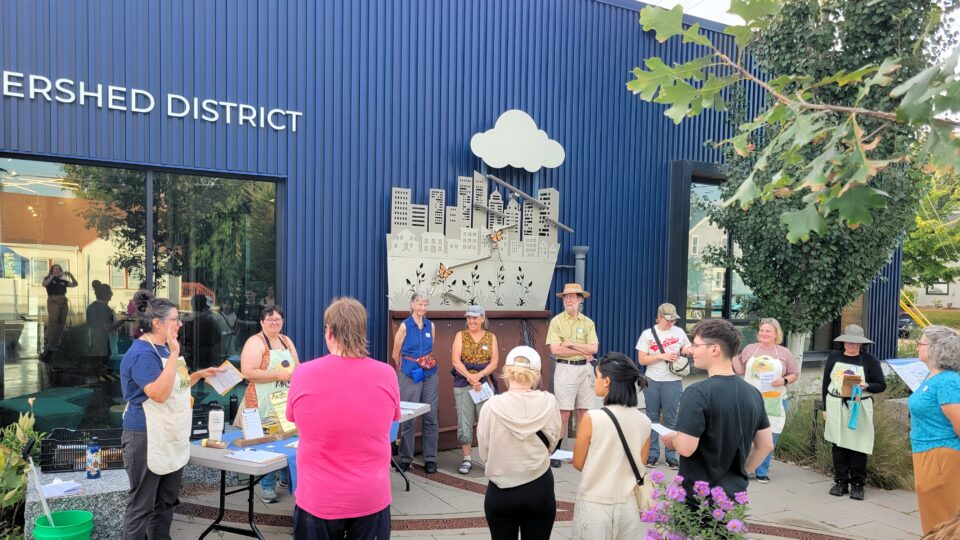 A group of people standing in a circle near a folding table outside of a blue-clad modern office building in early fall. They are listening to a person speaking.