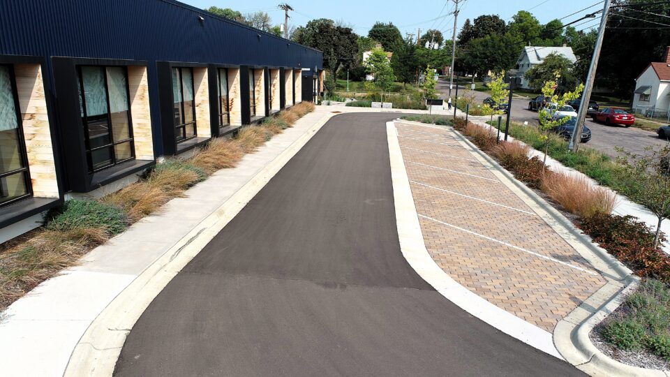 A black asphalt driveway along five angled parking spaces of red bricks. The lot is next to a modern blue-clad office building surrounded by low lanscaping and small trees in a residential neighborhood.