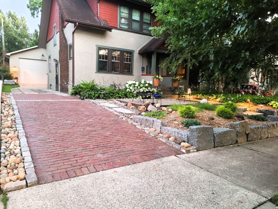 A red brick brick driveway along the side of a house with well-maintained landscaping, featuring neatly arranged stones and a variety of plants, including blooming white flowers. 