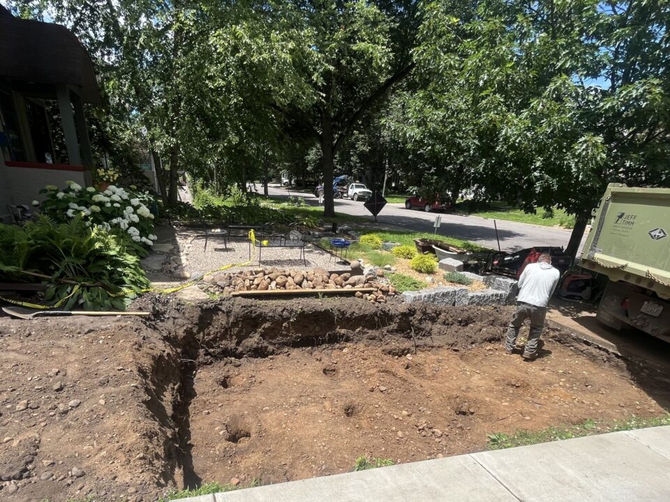 A person is standing next to a large, dug-out hole in a residential driveway. There are piles of dirt and rocks next to the hole. 